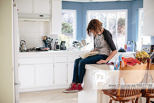 Teenager-Mädchen sitzt auf ihrem gekachelten Küchentisch und liest ein Buch