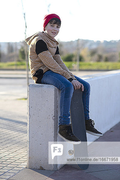 Junger Junge mit Kapuze sitzt mit seinem Skateboard auf einem Zaun