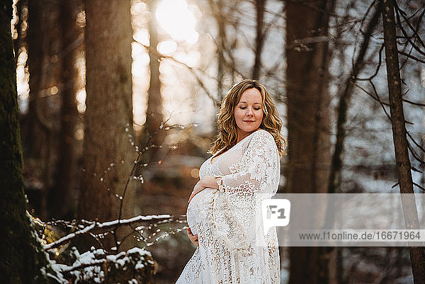 Schwangere Frau blickt im Winter bei goldenem Licht in den Wald hinunter