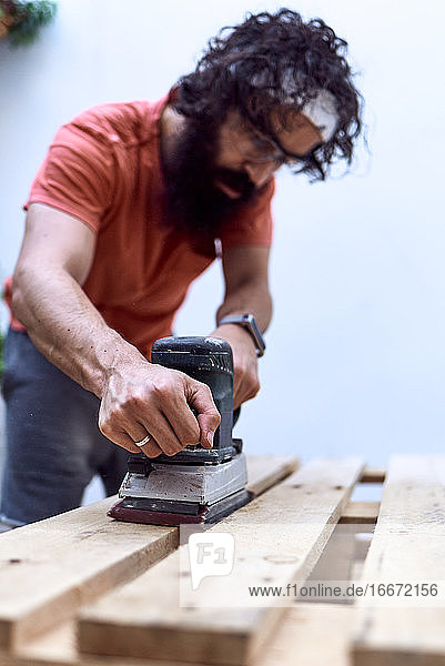 Junger Mann mit Bart  der ein Holzbrett mit einer Schleifmaschine poliert