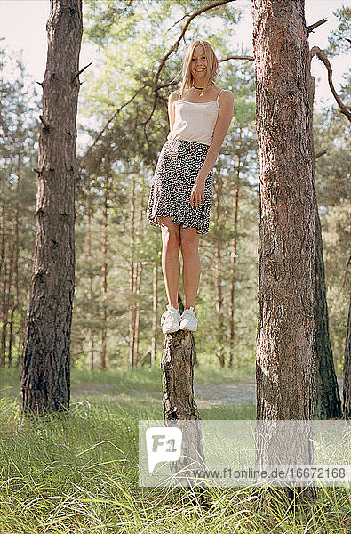 Blonde Frau im Rock steht auf einem Baumstamm im Frühlingswald.