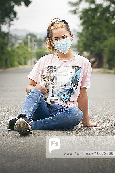 ein junges Mädchen oder eine Frau mit Gesichtsmaske sitzt auf der Straße und trägt eine junge weiß-graue Katze in einer verschwommenen natürlichen Umgebung
