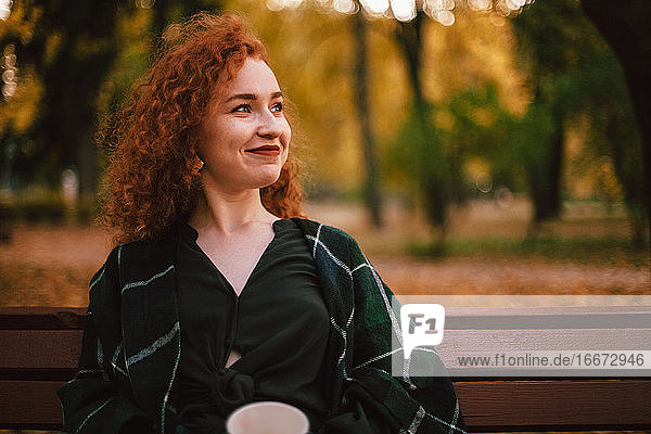 Portrait of smiling redhead woman sitting on bench in park in autumn