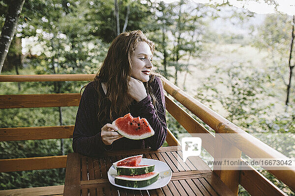 Junge rothaarige Frau isst Wassermelone auf dem Balkon eines Holzhauses