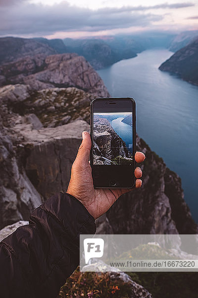 Hand hält Smartphone mit Bild der Szene im Hintergrund auf sie an norwegischen Fjorden.