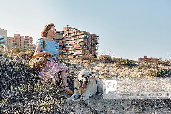 Frau schaut auf das Meer  während sie mit ihrem Hund im Sand sitzt