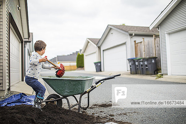 Young boy scooping pile of soil into wheelbarrow in back alley