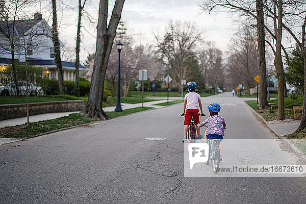 Rear view of two children biking together through neighborhood at dusk