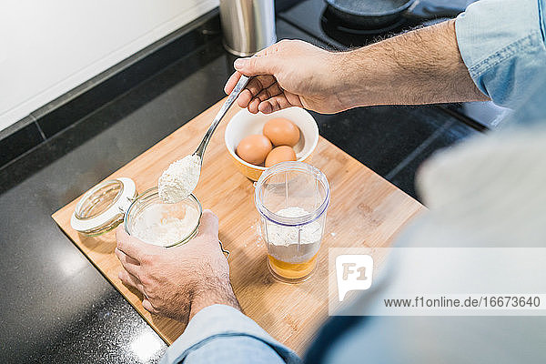 Mann im Jeanshemd beim Kochen in der Küche. Ein anonymer Mann hält einen Löffel mit Mehl