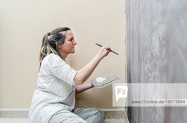 Eine blonde barfüßige Frau sitzt auf dem Fußboden und beginnt  eine braune Wand mit einem kleinen Pinsel grau zu streichen. Die Frau trägt ein weißes Hemd und eine graue Hose. Horizontales Foto.