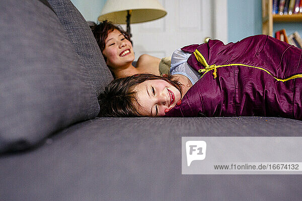 zwei glücklich lächelnde Kinder liegen in Schlafsäcken zusammen auf einer Couch
