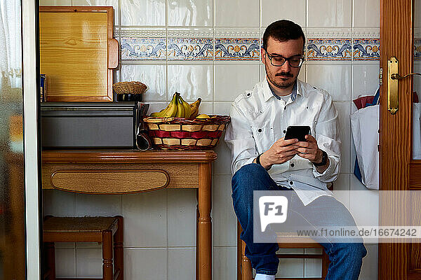 Junger Mann schaut auf sein Telefon  während er auf einem Küchenstuhl sitzt