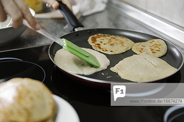 Crop person with spatula frying tasty pancakes on hot pan in kitchen
