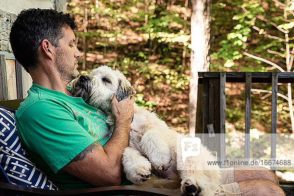 Man holding a fluffy dog on his lap outside on a deck in the woods.