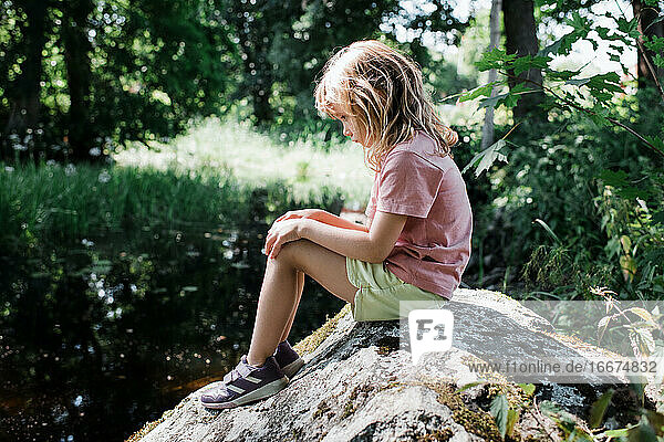 young girl looking sad and alone on a rock by a river in summer