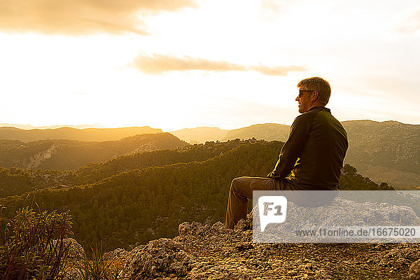 Ein Mann bei Sonnenuntergang auf einem Felsen sitzend mit Blick auf die Berge bei Sonnenuntergang