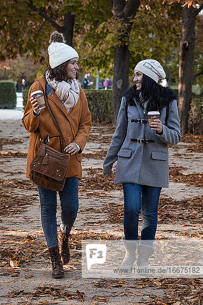 Beste Freunde plaudern im Herbst im Park mit ihrem Kaffee zum Mitnehmen. Sie tragen einen lässigen Mantel und eine Wollmütze.