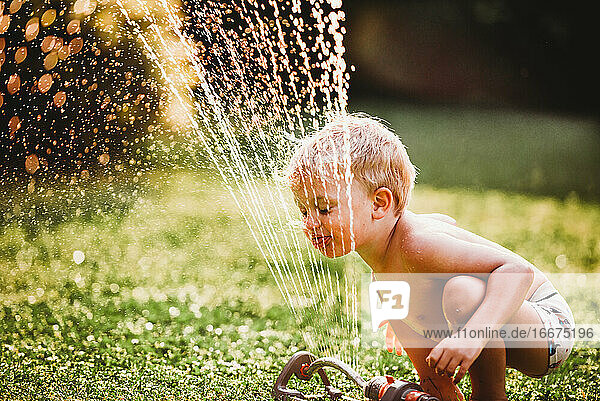 Kleiner Junge streckt die Zunge heraus und trinkt Wasser aus dem Rasensprenger im Garten