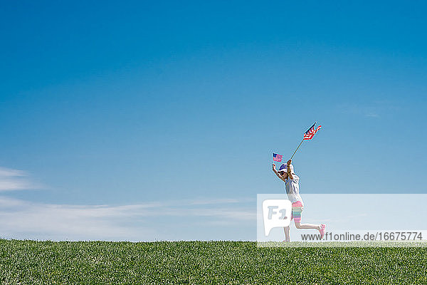 junges Mädchen springt über einen Grashügel und schwenkt 2 amerikanische Flaggen