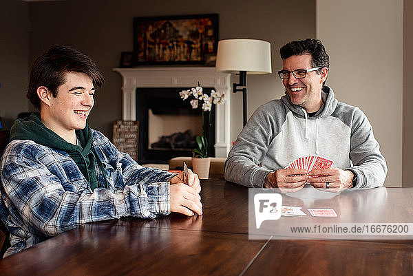 Vater und Sohn im Teenageralter lachen  während sie am Tisch Karten spielen.