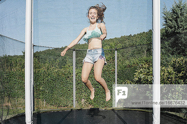 Horizontales Foto eines lächelnden jungen brünetten Teenagers  der auf einem Trampolin mit Netz auf dem grünen Hof im Freien springt. Das Mädchen trägt kurze Jeans und ein Top und sieht Spaß  aktiv