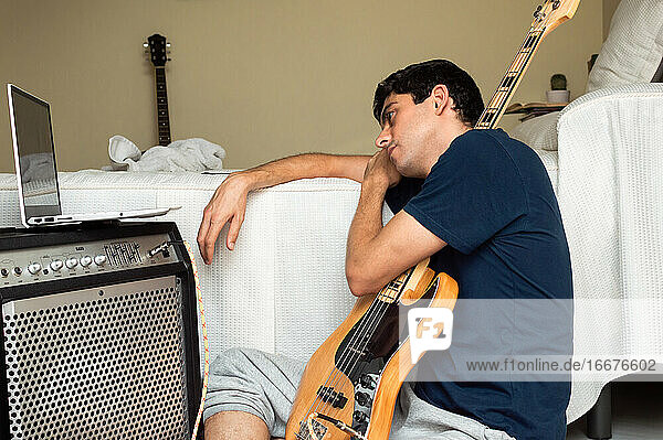 Gelangweilter Mann  der sich auf einem Laptop ein Tutorial ansieht  um zu Hause Bass spielen zu lernen.