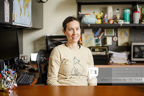 A smiling woman professor sits at her desk in an office