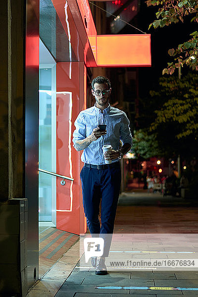Junger Geschäftsmann geht mit seinem Smartphone und seiner Kaffeetasse bei ni