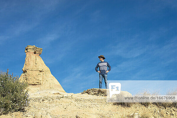 Vorderansicht eines Mannes mit Hut und Sonnenbrille  der auf Wüstendünen vor einem Felsgipfel steht