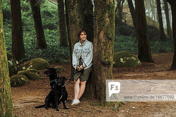 Frau mit zwei Hunden im Wald an einen moosbewachsenen Baum gelehnt