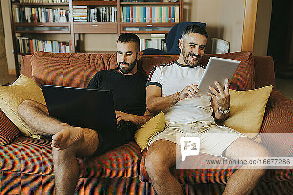 schwules Paar auf dem Sofa sitzend mit Laptop und Tablet