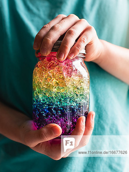 Nahaufnahme von Kinderhänden  die ein Glas mit regenbogenfarbenen Kristallen halten.