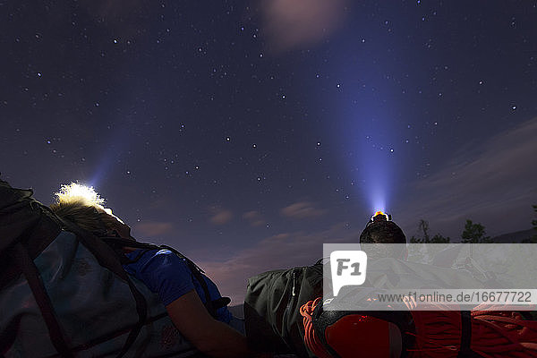 zwei Personen  die sich unter den Sternen mit Scheinwerfern auf einen Rucksack legen