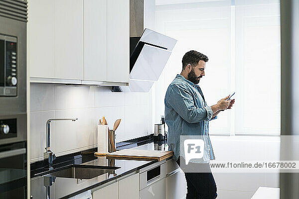 Man cooking crepes in the kitchen with a mobile phone in a denim shirt