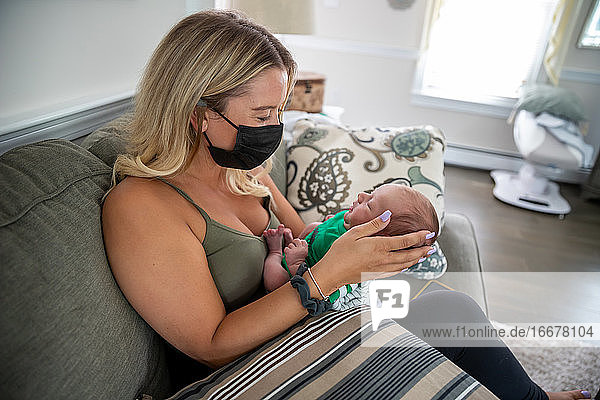 Blonde Frau mit Gesichtsmaske hält ein schläfriges Neugeborenes.