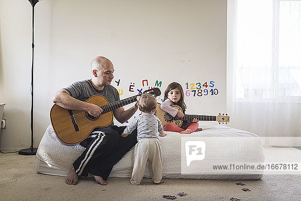Dad and daughter playing guitar in kid's room with 1 yr old watching