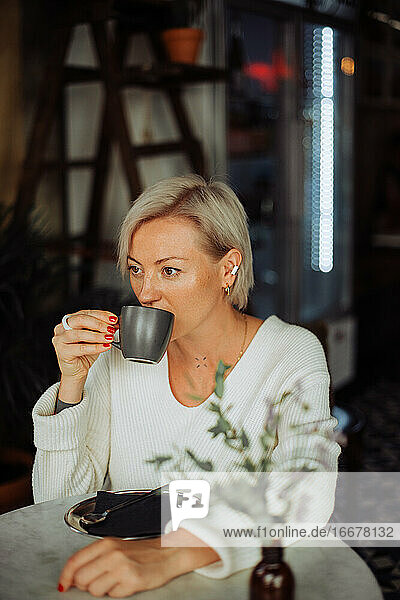 Stylish blond woman drinking coffee while sitting at table in cafe