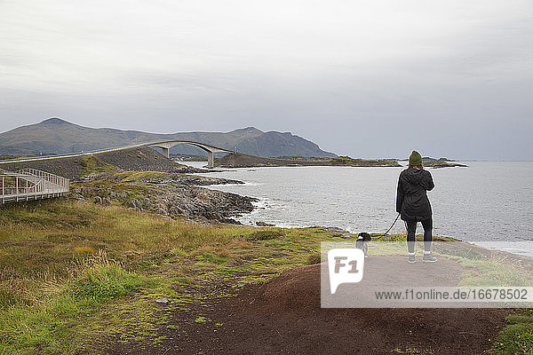 Eine Frau steht mit ihrem Hund auf einem grasbewachsenen Hügel am Meer in Norwegen