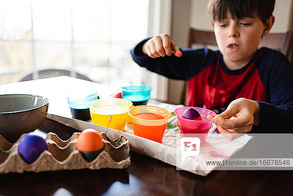 Ein Junge taucht ein Ei in eine Schüssel mit Farbstoff  um es für Ostern zu färben.