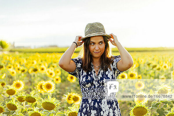 Young attractive brunette woman posing in her designer dress in a field of sunflowers and wearing a hat