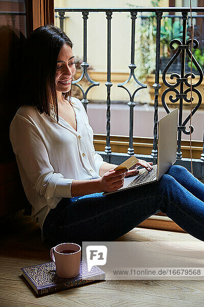 Junge Frau kauft ein und bezahlt online auf einem Laptop mit Kreditkarte  während sie sich zu Hause am Fenster entspannt.