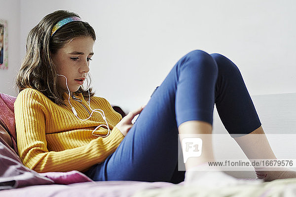 Mädchen liegt auf dem Bett und schaut auf ein Tablet und trägt Kopfhörer