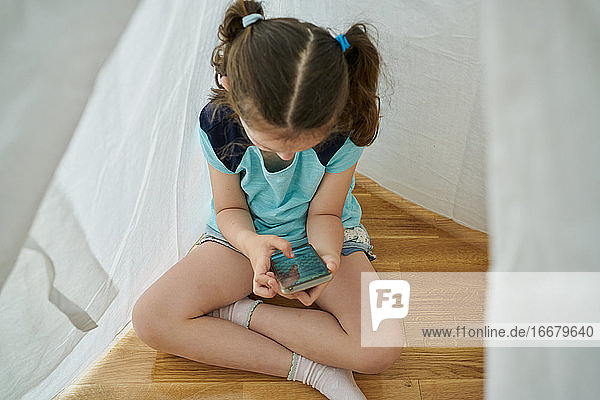 Kleines Mädchen schaut auf ihr Smartphone in einem weißen Tipi-Zelt in ihrem Haus. Technologie-Konzept