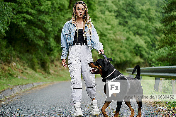 Junge Frau mit blondem geflochtenem Haar  Jeansjacke und weißer Jeans  die an einem regnerischen Tag mit ihrem Hund spazieren geht