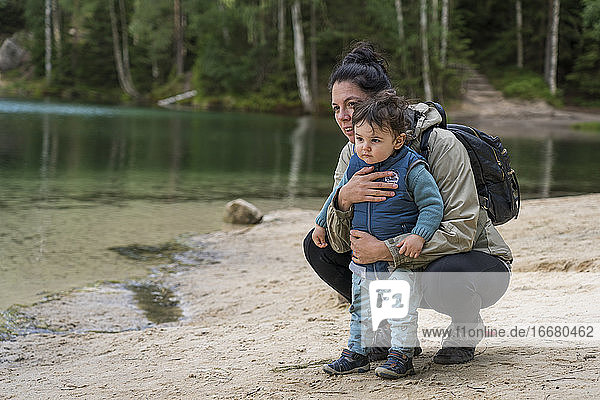 Mother with baby boy looking away at lakeshore while hiking in f