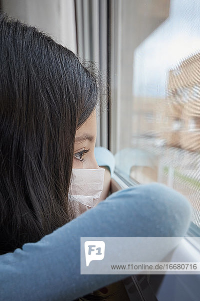 Girl with face mask looking out the window quarantined by coronavirus