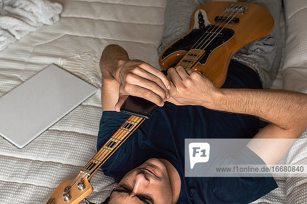 Ein Mann liegt auf einem Bett und benutzt ein Mobiltelefon  nachdem er E-Bass gespielt hat.