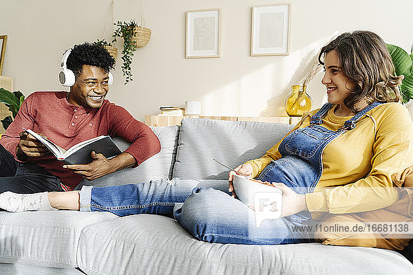 Pregnant wife eating on the couch with her husband who is listening to music and reading a book. Interracial couple concept