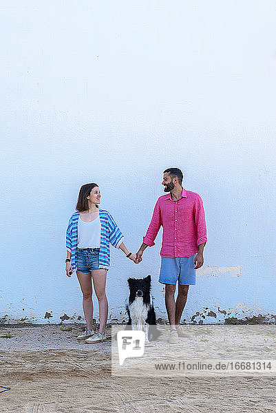 Glückliches Paar steht mit Hund an der Wand und hält sich an den Händen