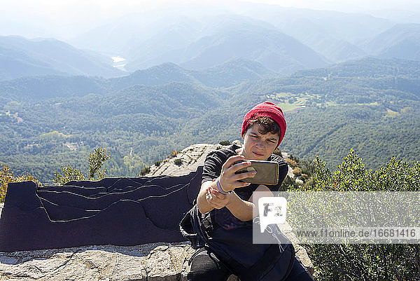 Junger Mann mit roter Wollmütze sitzt am Berg und macht ein Selfie
